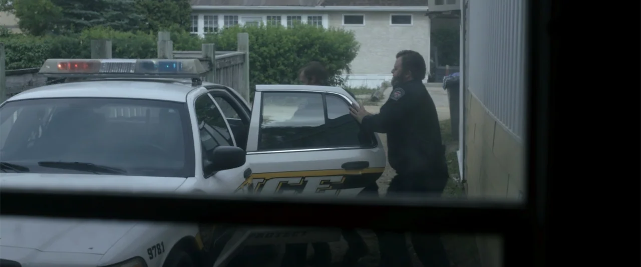 Radius (2017) movie still: medium — A residential driveway with a white patrol car parked next to a house siding. A police officer is assisting a…