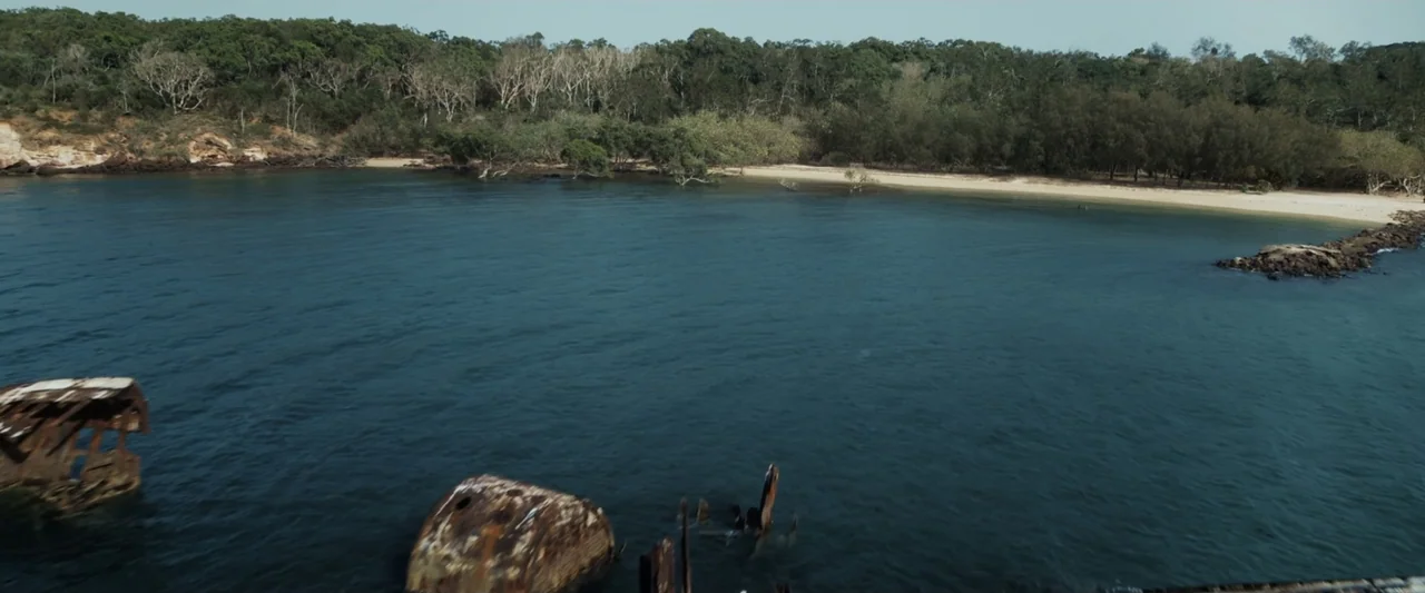 Great White (1998) movie still: establishing — A secluded coastal inlet with sandy beaches surrounded by dense trees and shrubs. Two rusted metal…