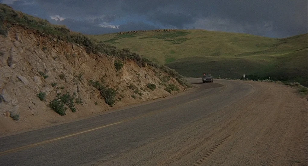 Vanishing Point (1971) movie still: wide — A rural asphalt road curves through rolling, grassy hills under a dark, storm-filled sky. A small,…