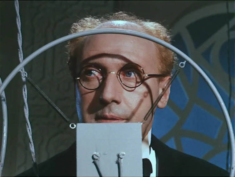 All These Women (1964) movie still: closeup — A man stands in a dimly lit, blue-hued laboratory environment behind a metal construction. He stares…