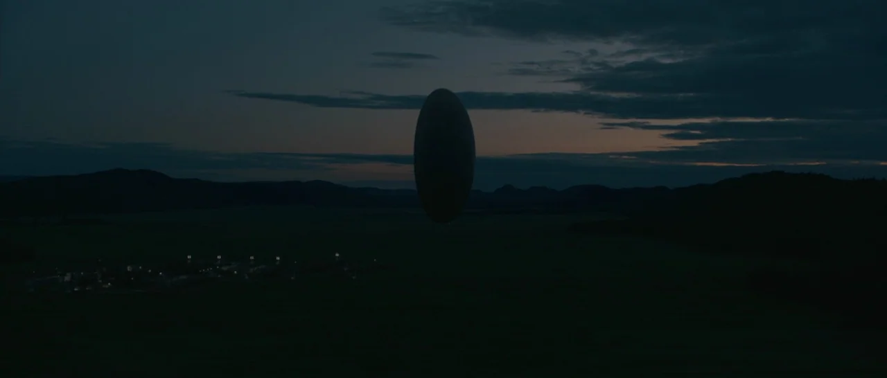 Arrival (2016) movie still: establishing — A vast, dark rural field is set against a twilight sky with low mountain silhouettes. A large monolithic…