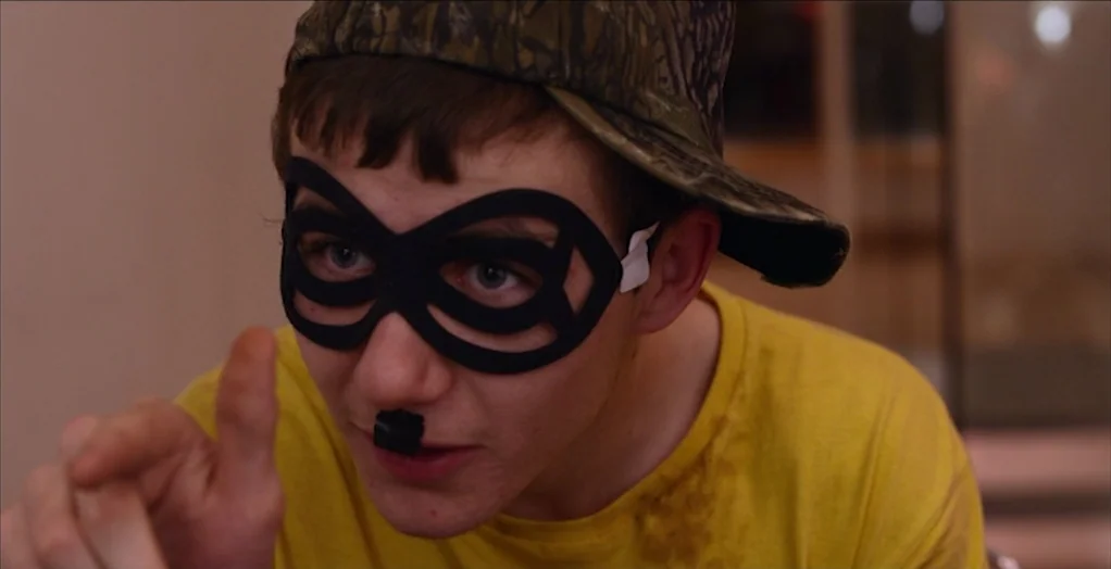 Dollhouse (2012) movie still: closeup — A close-up view of a young person wearing a mask and cap. He points forward while dressed in a yellow t-shirt.