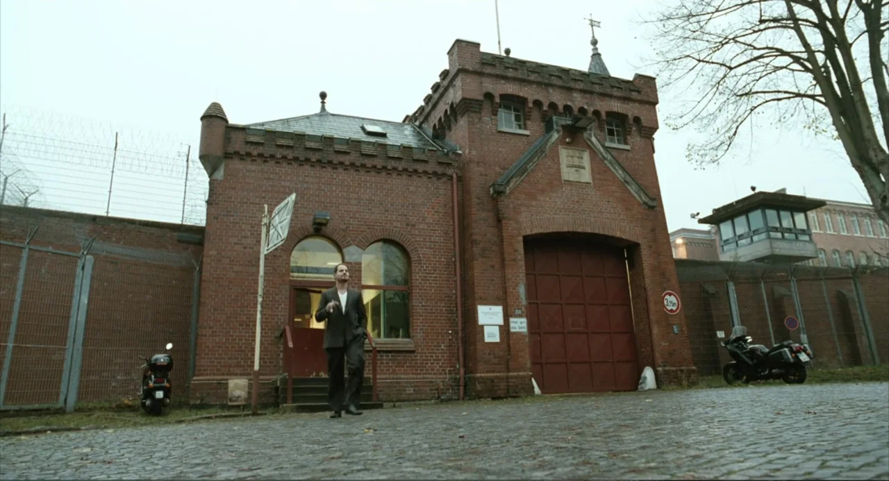 Soul Kitchen (2009) movie still: wide — A red brick prison exterior with a large arched gate and perimeter barbed wire. A man in a dark suit walks…