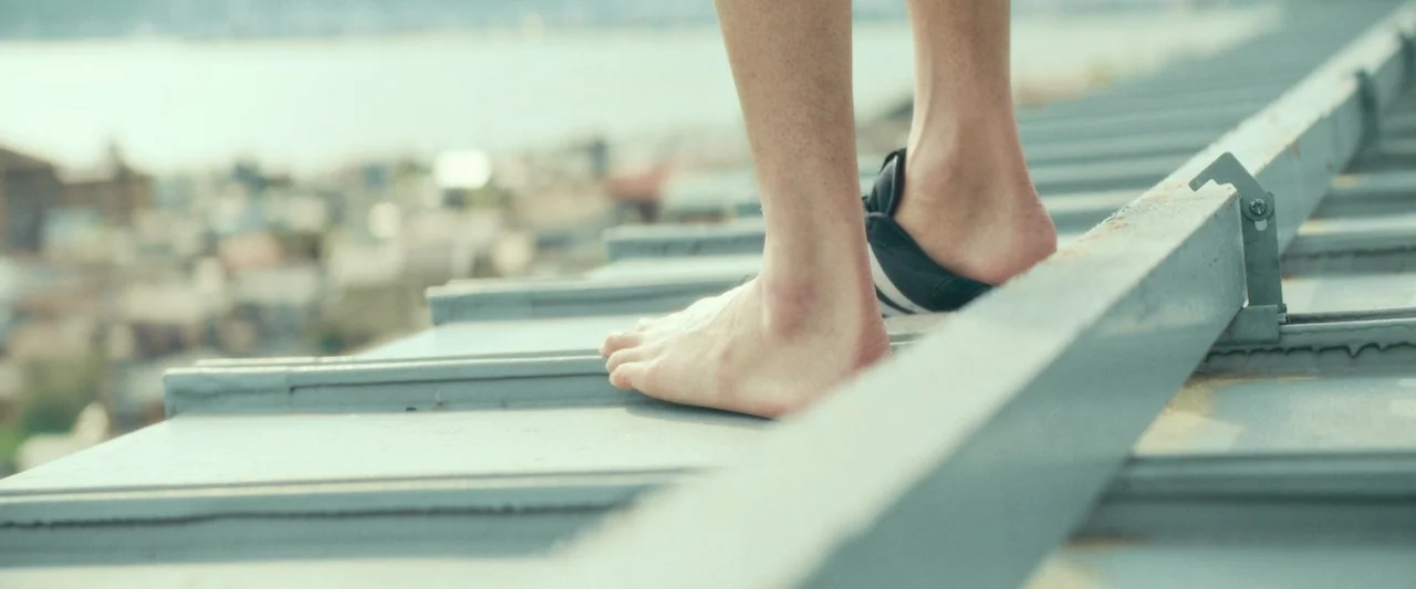 Monster (2023) movie still: closeup — A person stands barefoot on the corrugated metal edge of a rooftop. The blurry urban landscape is visible in…