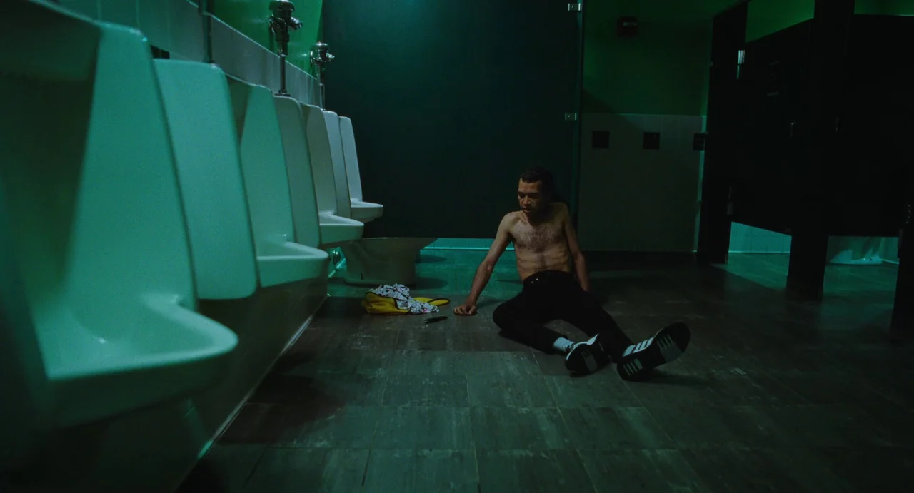 I Saw The TV Glow (2024) movie still: fullbody — A public restroom with rows of white urinals and green walls. A shirtless man sits on the floor…