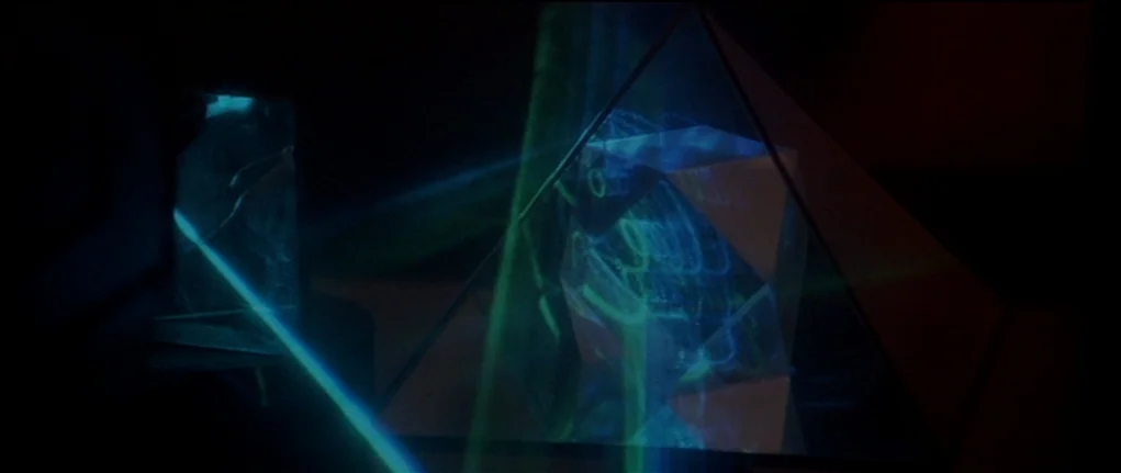 Demon Seed (1977) movie still: closeup — A dark space contains glass geometric shapes illuminated by concentrated light beams. The lasers create…
