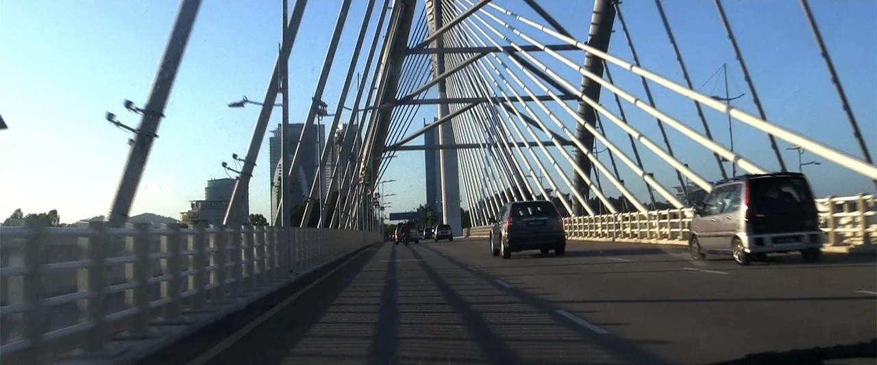 The Kleptocrats (2018) movie still: wide — A cable-stayed bridge spans across a wide roadway with tall concrete support towers. Several vehicles…
