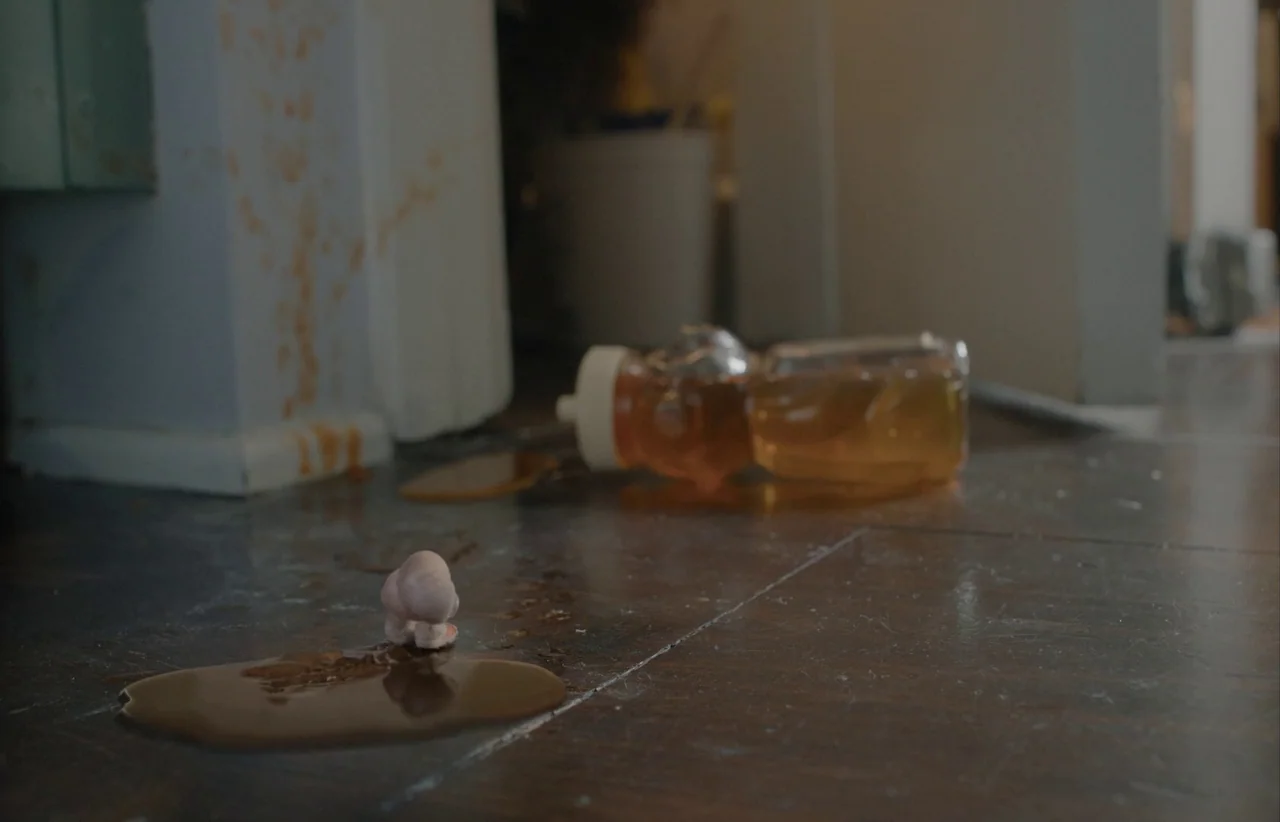 Marcel the Shell with Shoes On (2021) movie still: closeup — A dark tiled floor features a spilled plastic honey bottle and a small figurine.…