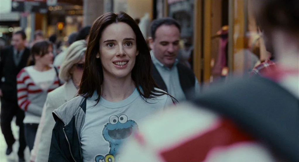 Sidewalls (2011) movie still: medium — A crowded urban sidewalk filled with pedestrians walking past storefronts. A young woman walks through the…