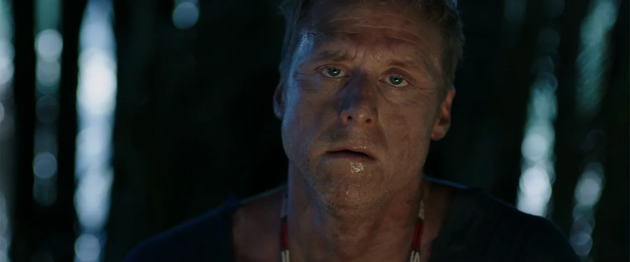 Playing God (2021) movie still: closeup — A dense, dark jungle environment at night with blurred foliage in the background. A man stands in the…