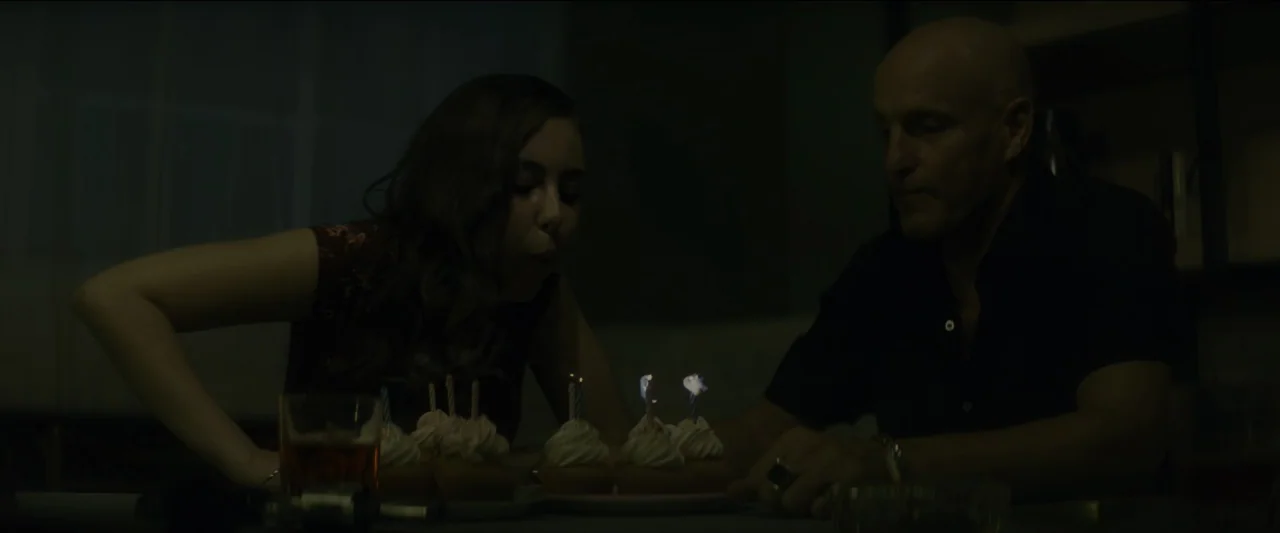 Kate (2021) movie still: medium — A dimly lit room holds a table with a plate of small frosted cupcakes. A woman leans forward to blow out the…