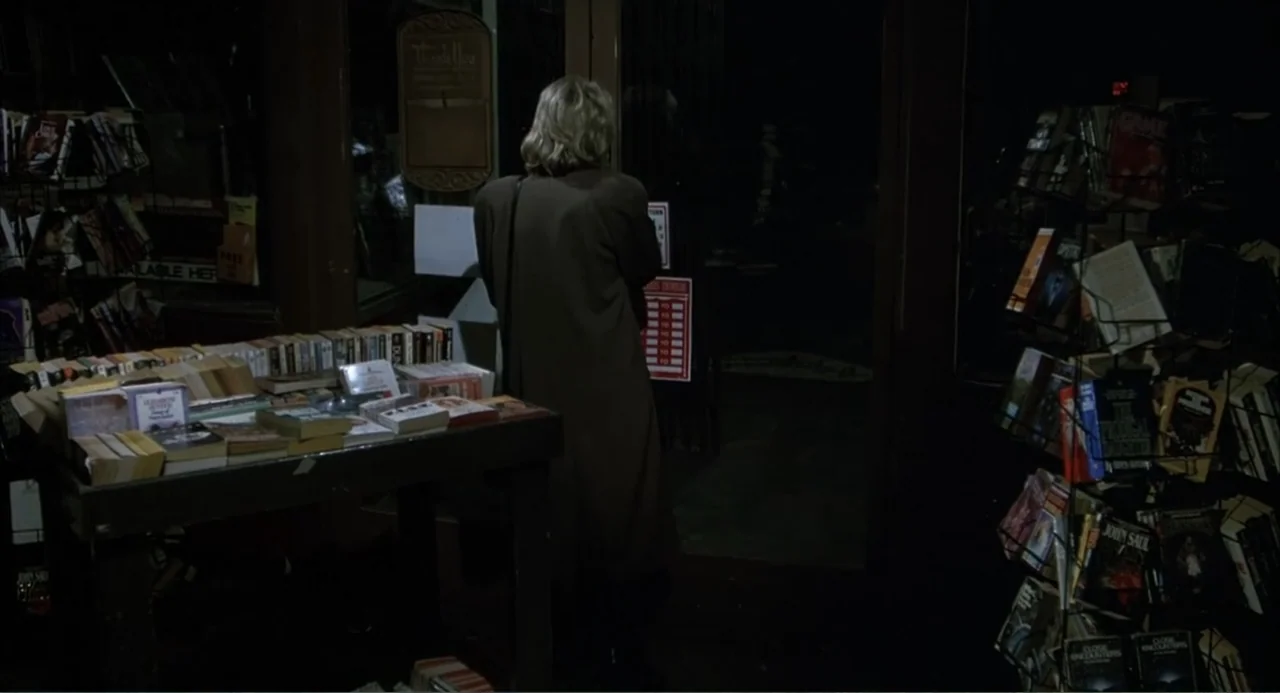 I, Madman (1989) movie still: medium — A dimly lit retail store with shelves packed with books and paperback magazines. A blonde woman in a long…