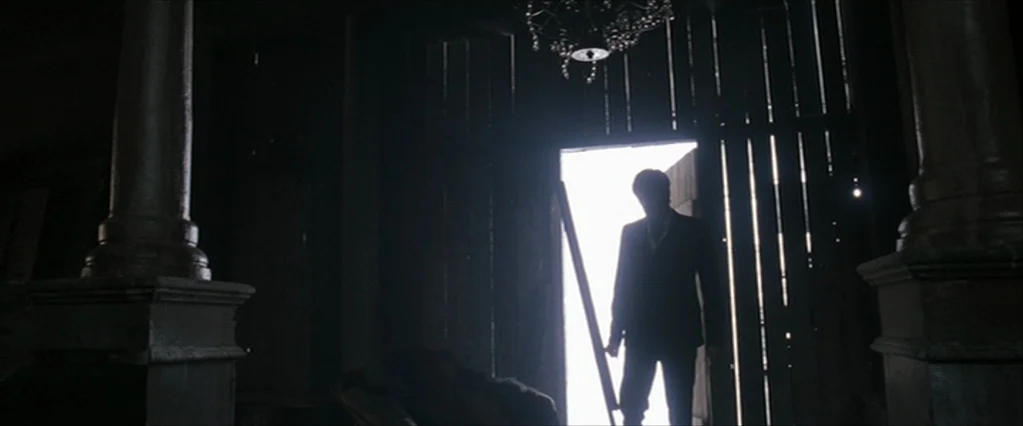 The Brothers Bloom (2008) movie still: fullbody — A dark, dilapidated interior space with visible support pillars and a low-hanging chandelier. A…