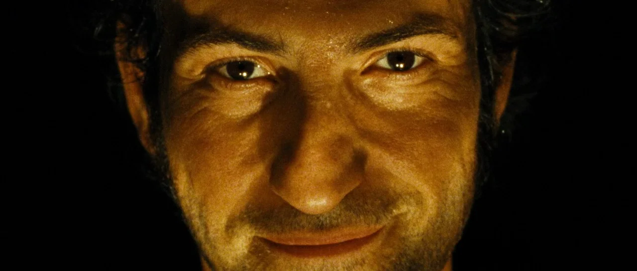 Let The Corpses Tan (2017) movie still: closeup — A man's face is illuminated from below against a pitch black background. The subject stares…