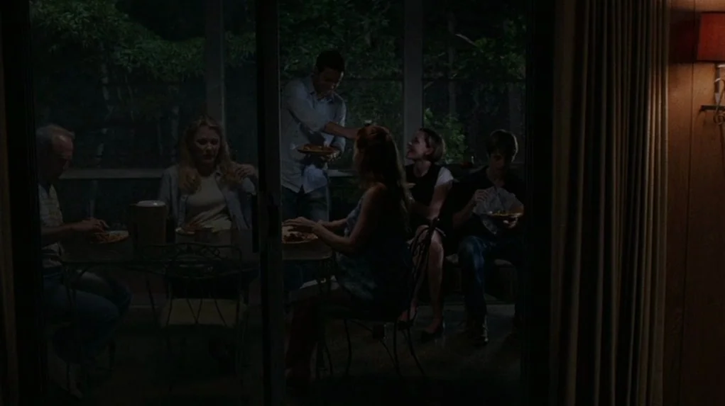 Junebug (2005) movie still: wide — A group of six people eat dinner on a dimly lit screened-in wooden porch. They are seated at a round metal table…