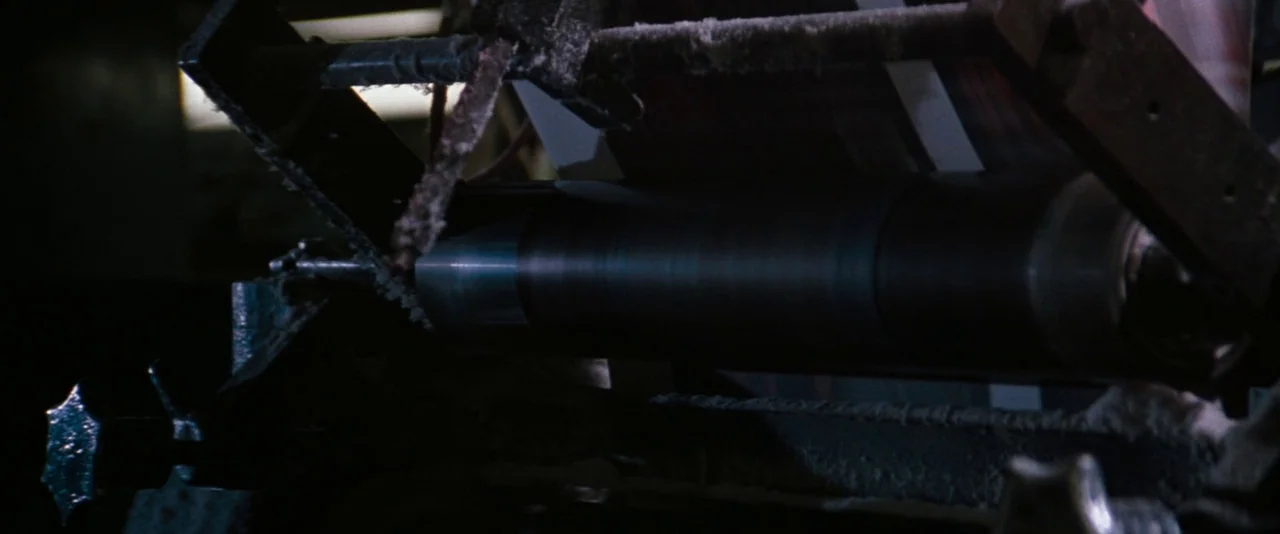 In The Mouth of Madness (1994) movie still: closeup — A close-up view of a heavy mechanical industrial machine with rotating rollers. The metallic…