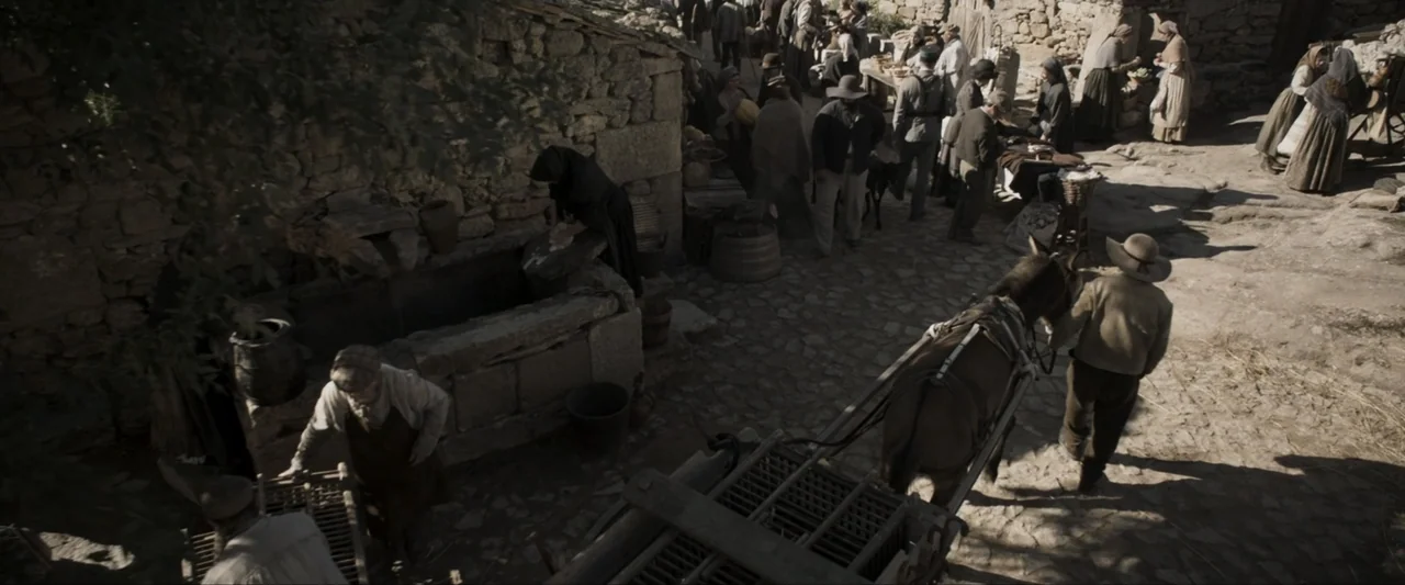 Fatima (2020) movie still: wide — A stone-walled village square shows a busy outdoor market with multiple inhabitants. Peasants walk among stalls…