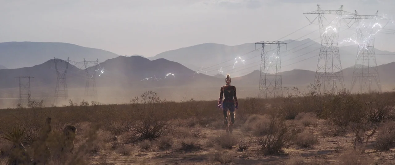 Captain Marvel (2019) movie still: wide — A vast, dry desert landscape features distant mountains and multiple large metal utility towers. A single…