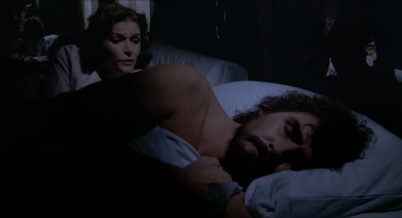 The Amityville Horror (1979) movie still: medium — A dimly lit bedroom features a woman watching a sleeping man in a bed. The man is covered by…