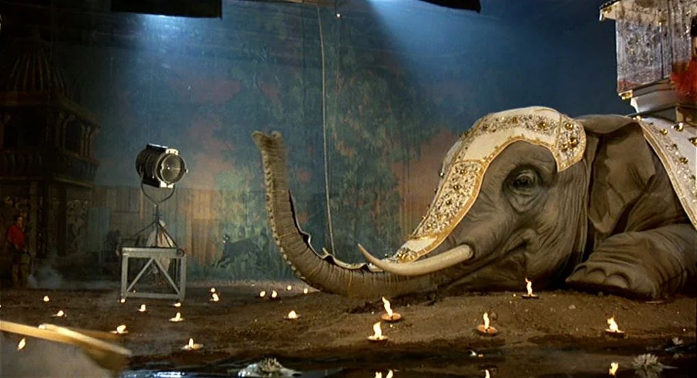 Intervista (1987) movie still: wide — A large indoor sound stage features a painted jungle backdrop and a mechanical elephant. Numerous small lit…