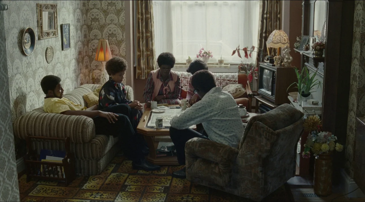 Red, White & Blue (2020) movie still: wide — A 1970s living room features patterned wallpaper and a retro carpet underfoot. Four individuals sit on…