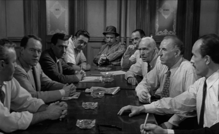 12 Angry Men (1957) movie still: wide — A group of twelve men sits around a long wooden table in a room. They are engaged in a discussion while…