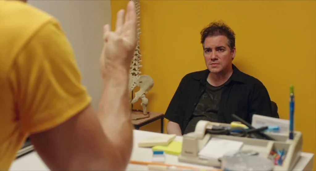 Results (2015) movie still: medium — An office with a bright yellow wall features a skeletal model on a stand. A man wearing a black shirt sits…