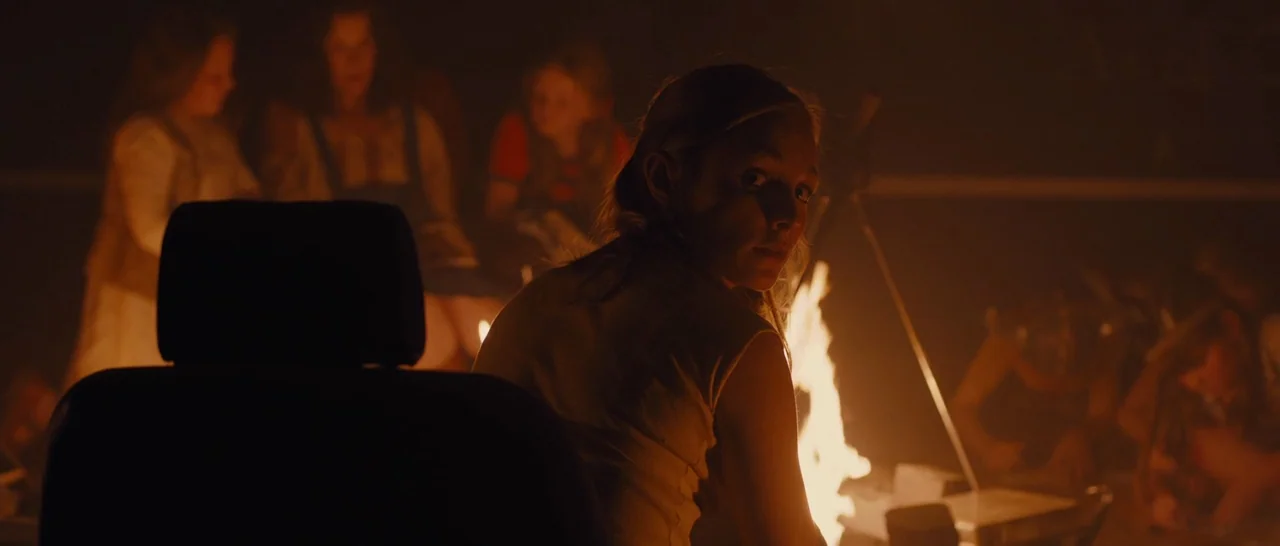 High-Rise (2015) movie still: medium — A group of people sits around an open fire in the dark night. A woman in the foreground looks back over her…