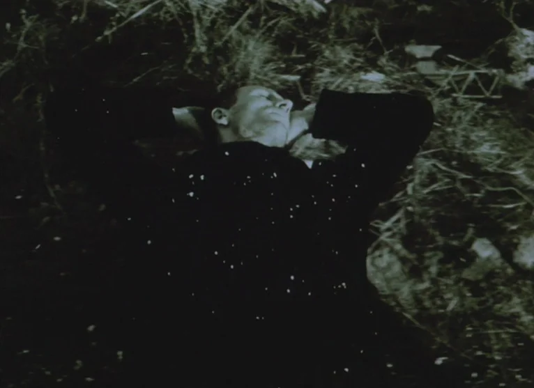 A Visitor to a Museum (1989) movie still: medium — An outdoor field is covered in dry grass and vegetation. A man lies on his back with hands behind…