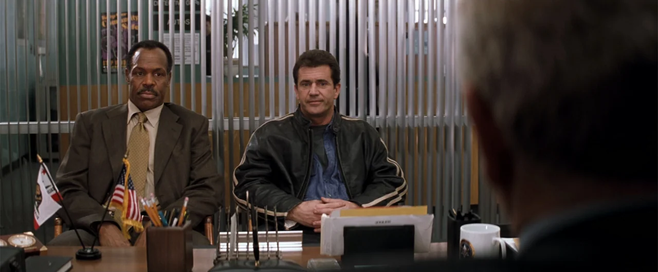 Lethal Weapon 4 (1998) movie still: medium — A modern police office features metallic vertical blinds and a desk with office supplies. Two men sit…