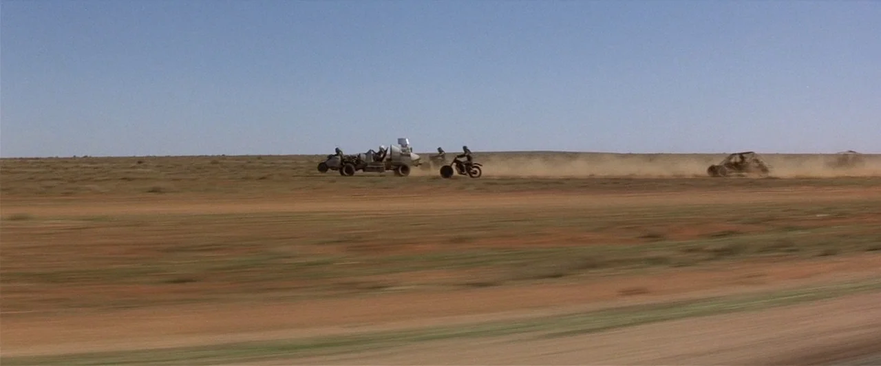 Mad Max 2 (1981) movie still: wide — An expansive, flat, and arid desert plain stretches toward a distant, low horizon line. A group of modified…