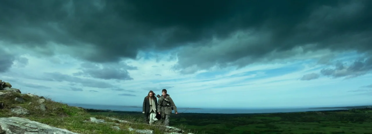 28 Years Later (2025) movie still: wide — A grassy, elevated landscape overlooks a distant coastline under a thick, overcast sky. Two figures trek…