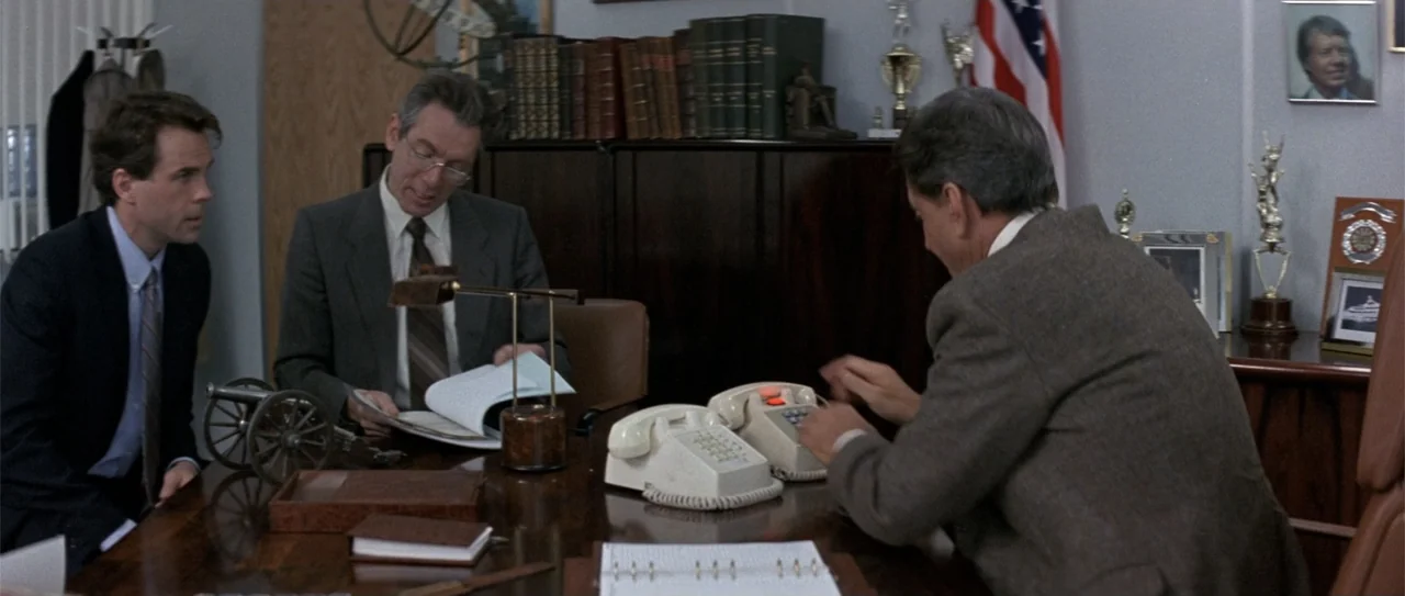 The Russia House (1990) movie still: medium — A professional office contains bookshelves, a dark wooden desk, and a wall-mounted American flag.…