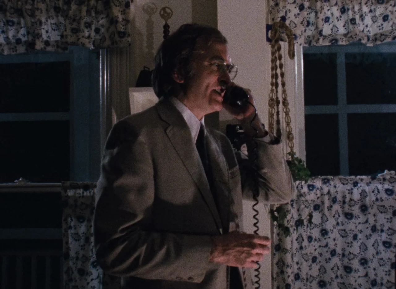 Basket Case (1982) movie still: medium — A residential room features dark windows covered by floral-patterned curtains at night. An older man in a…