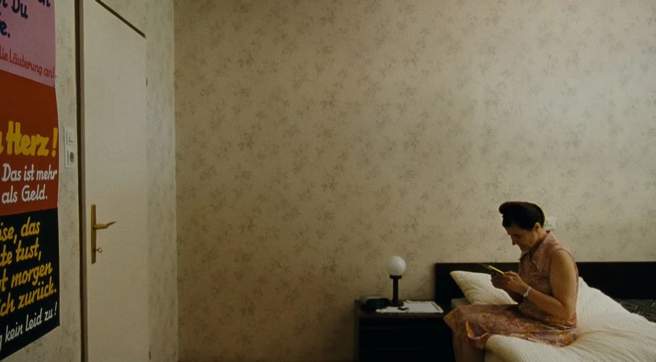 Paradise: Faith (2012) movie still: medium — A spare hotel room features floral wallpaper and a bed against a blank wall. An older woman sits on the…