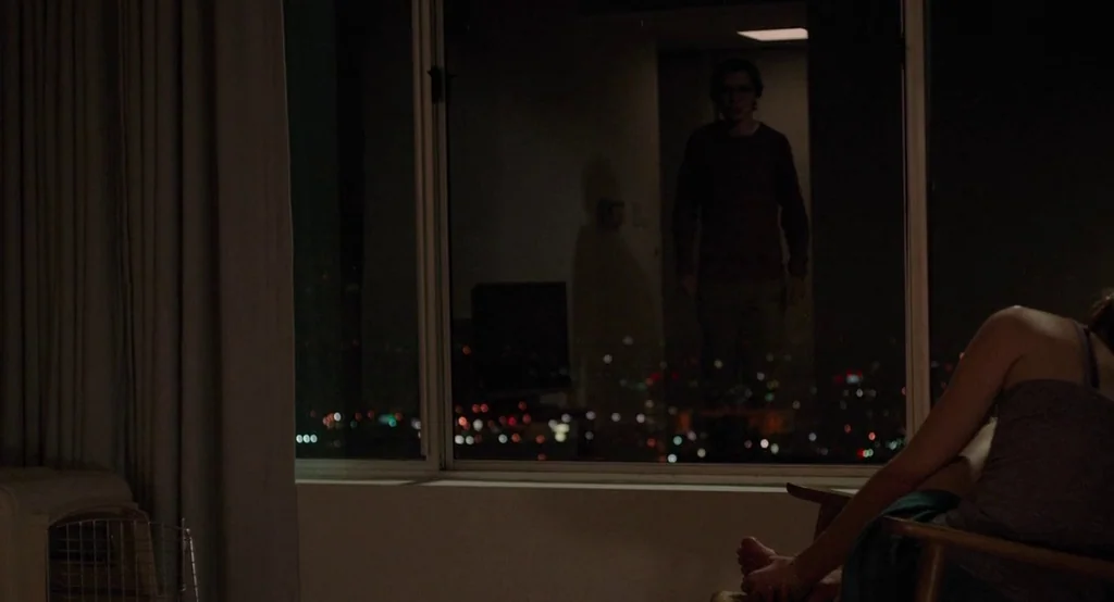 Ruby Sparks (2012) movie still: medium — A dark apartment room looks out onto a blurry city skyline at night. A woman sits in a chair in the…