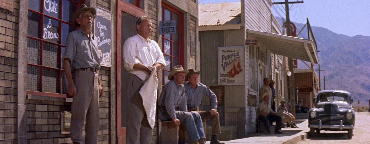 Bad Day At Black Rock (1955) movie still: wide — A dusty, sun-drenched row of wooden storefronts lines a dirt-road town under a mountain. A group of…