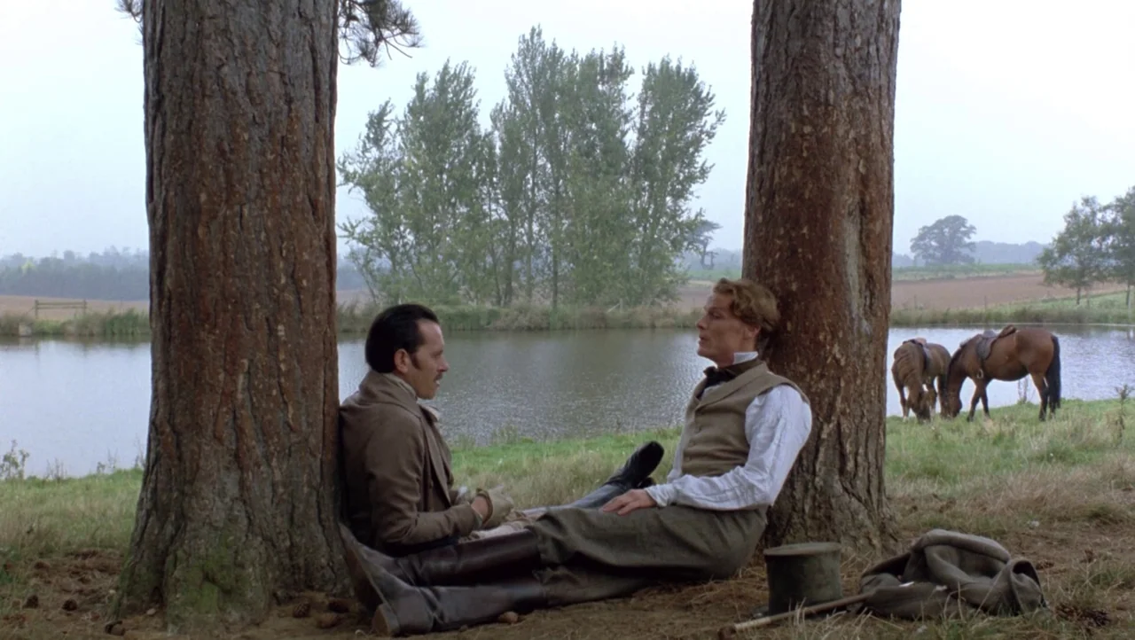 Mountains of the Moon (1990) movie still: fullbody — Two men in nineteenth-century attire sit against two large trees in a grassy field. A small…