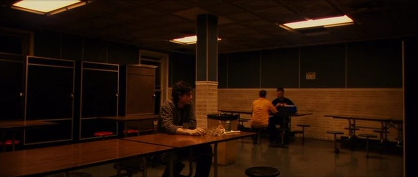 Bubble (2005) movie still: wide — A dimly lit cafeteria space contains rows of metal tables and dark wall cabinets. One man sits alone in the…