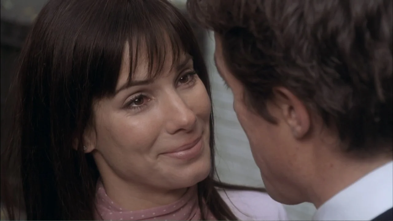 Two Weeks Notice (2002) movie still: closeup — An interior space with a man and woman positioned close together. The woman smiles at the man who is…