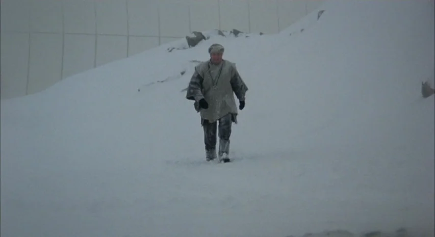 Quintet (1979) movie still: fullbody — A vast, flat, snow-covered landscape stretches toward a distant, barely visible wall. A man in a heavy grey…