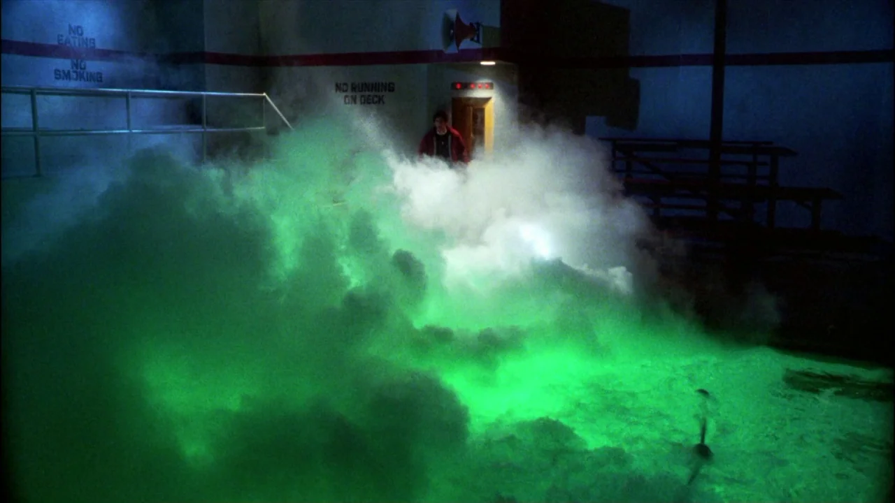 Gremlins (1984) movie still: wide — An indoor pool area features painted warning signs on walls and tiered metal seating. A young man stands at the…