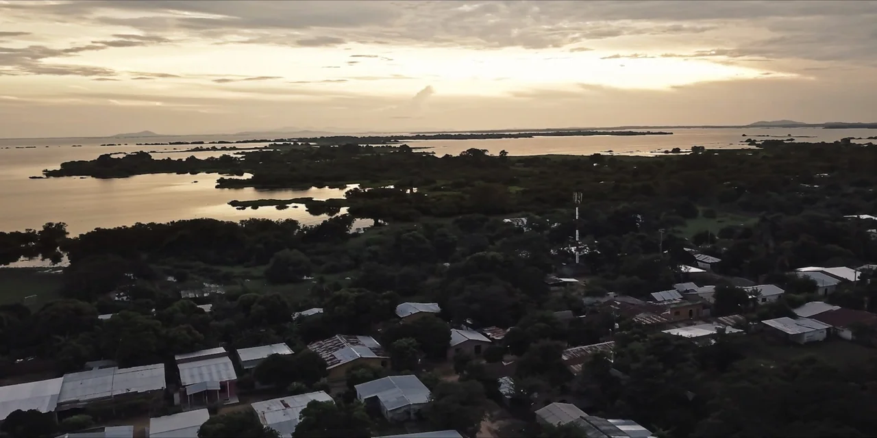Leyenda Viva (2022) movie still: establishing — A rural village with metal-roofed houses sits alongside a expansive lake at sunset. Lush green…