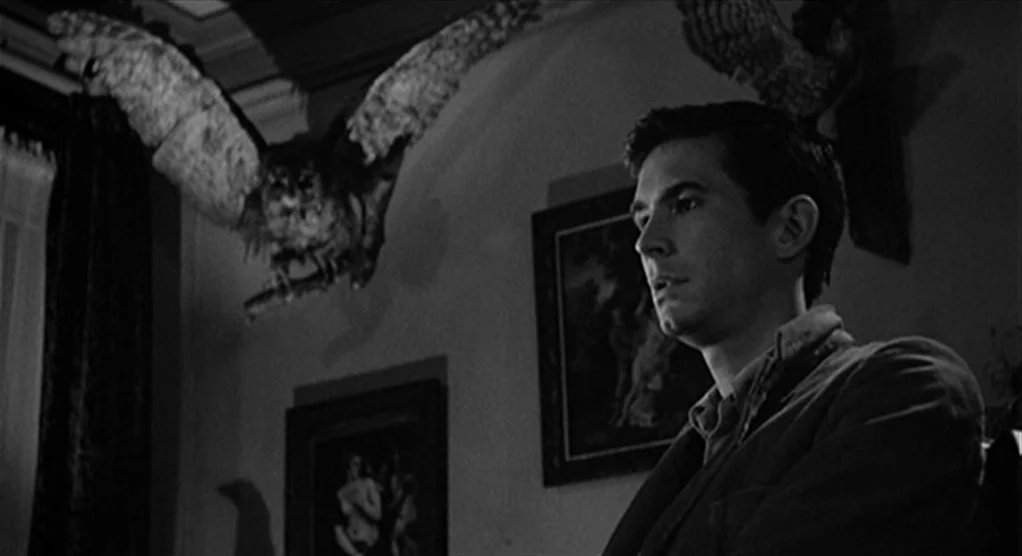 Psycho movie still: medium — A dimly lit room features walls decorated with framed paintings and taxidermy birds. A young man stands in the…