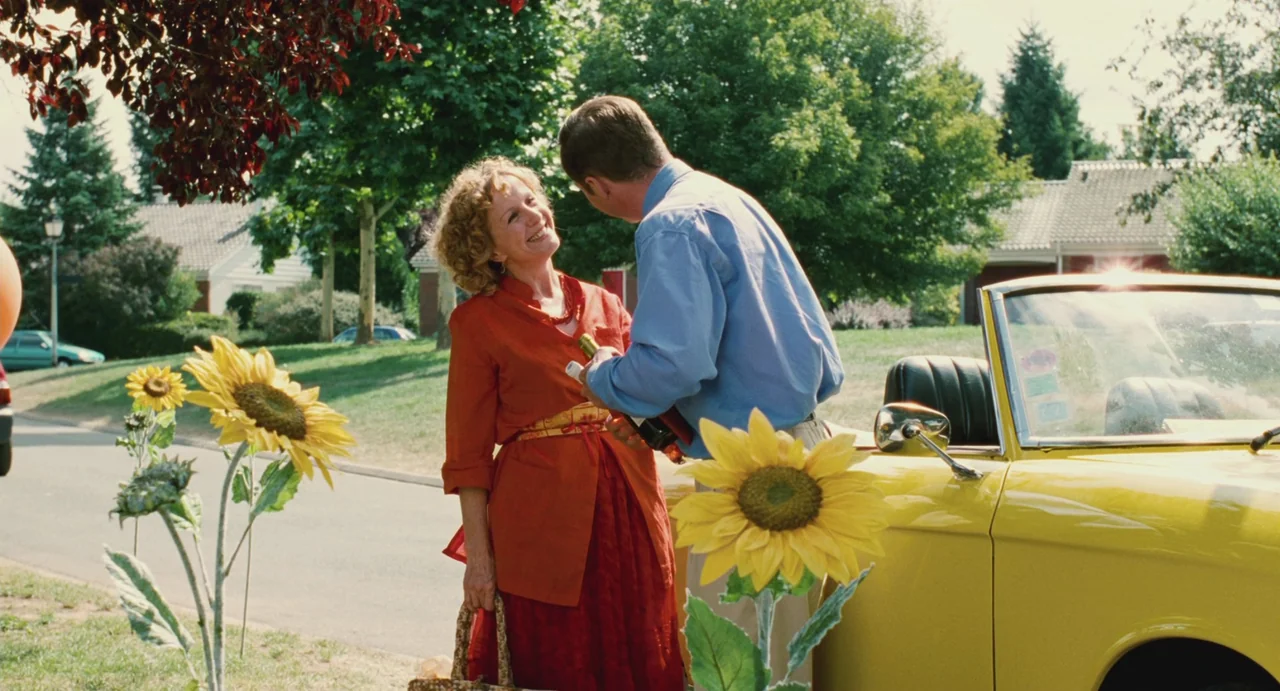 Ma Vie En Rose (1997) movie still: medium — A suburban street features a yellow convertible parked next to oversized artificial sunflowers. A woman…