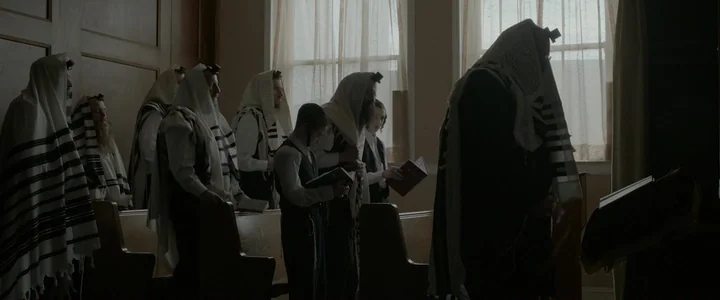 To Dust - Nightmares (2018) movie still: wide — A row of men wearing tallitot stand inside a dimly lit synagogue interior. They hold small prayer…