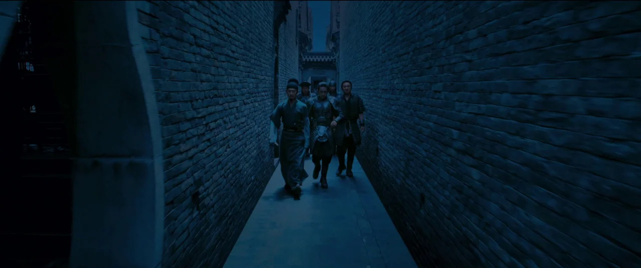 Full River Red (2023) movie still: wide — A narrow corridor lined with dark grey brick walls and stone flooring. A group of men walk forward in a…