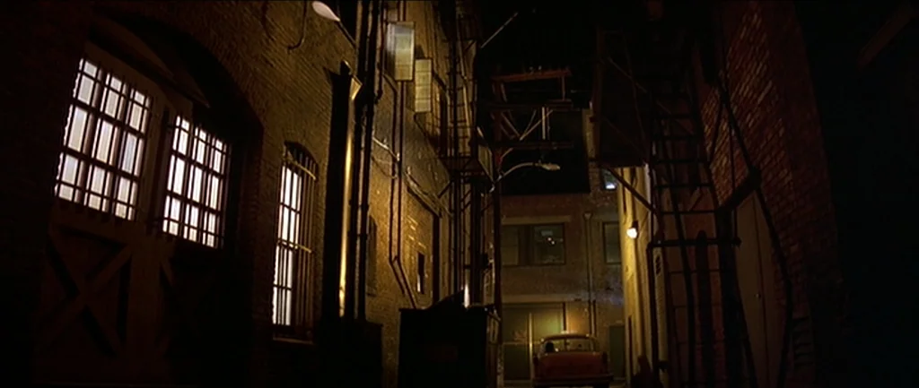Pulp Fiction (1994) movie still: establishing — A narrow city alley is framed by dark red brick walls and metal fire escapes. A parked taxi is…