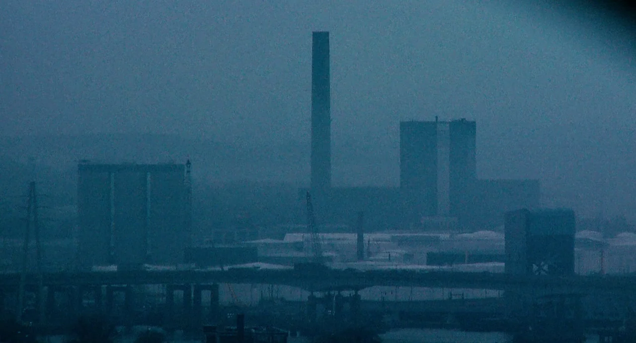 Another Earth (2011) movie still: establishing — A dark industrial landscape features multiple large factory buildings under a thick blue fog. A…