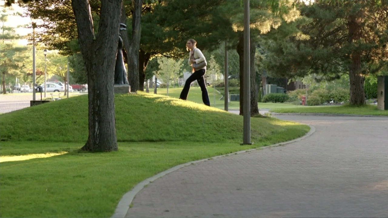 At Middleton (2013) movie still: wide — A park landscape features a curved paved walkway alongside a grassy hill with mature trees. A young woman…